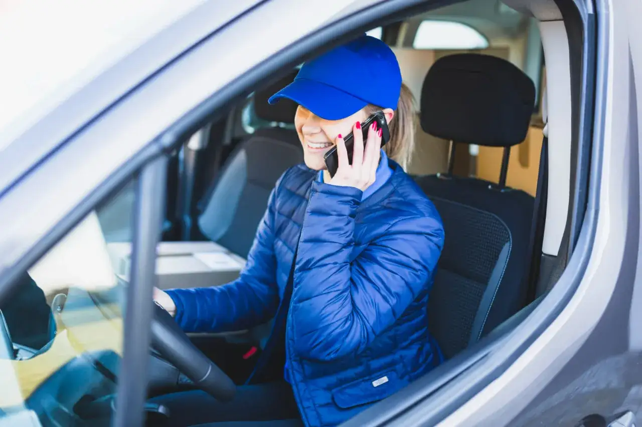 Uśmiechnięta kurierka w niebieskiej czapce i kurtce rozmawia przez telefon, siedząc za kierownicą. Dowozi paczki do paczkomatów InPost.