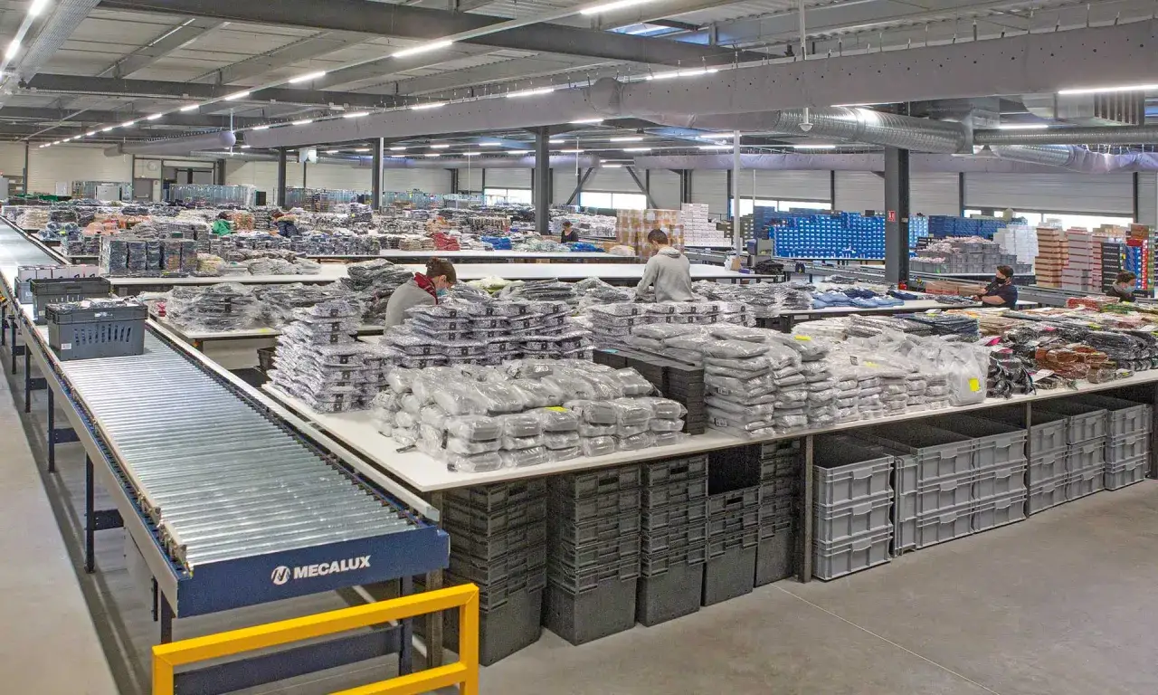 Pracownicy sortują odzież w magazynie. Widać taśmociąg, stoły z zapakowanymi produktami i pojemniki. To pokazuje różne funkcje magazynu.